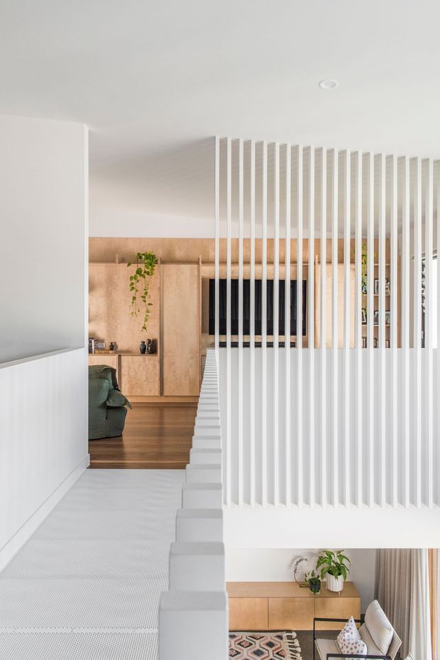 Bright contemporary interior showing a white staircase and landing screened by closely spaced vertical white battens. Beyond the screen is a warm timber-clad living area with built-in cabinetry, wall-mounted television, greenery and a partially visible sofa. The composition also includes a small inset view of the seating area at the lower right. Clean lines, pale surfaces and warm timber create a refined, open-plan feel.