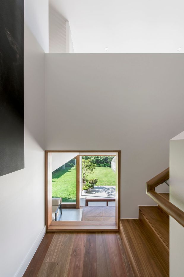 A bright contemporary stairway or landing viewed from above, with warm timber flooring and timber stair treads contrasted against smooth white walls and a large dark feature panel. A timber-framed glazed opening at the bottom of the stairs looks onto a garden and adjoining living area. The space has a high ceiling, minimalist detailing, and strong natural light.