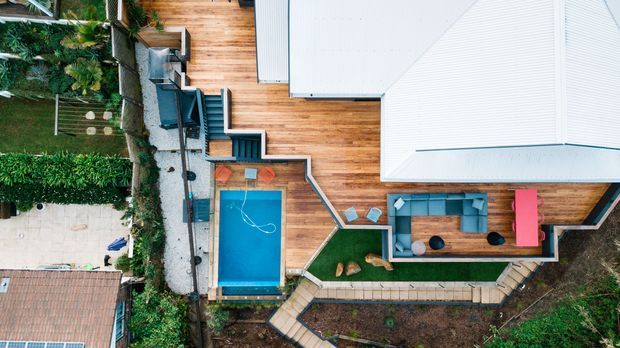 Aerial view of a contemporary residential property with multiple white metal roof volumes, extensive timber decking, a bright blue swimming pool, outdoor seating, landscaped garden beds, and a small lawn area. The image emphasizes the building footprint, outdoor living areas, and pool layout.