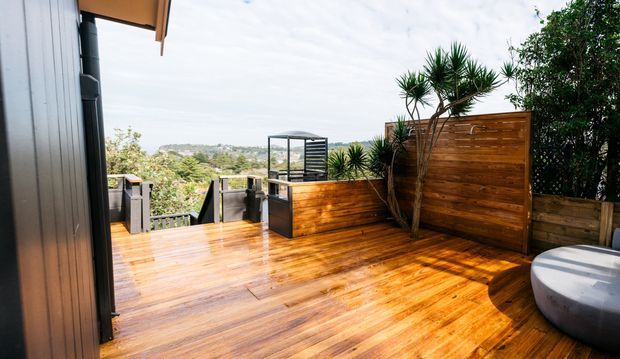 A sunlit timber outdoor deck and entertaining area beside a contemporary home, featuring warm hardwood decking, matching privacy screens and fencing, dark metal-framed elements, planted yucca-like trees, and a partially visible round spa or plunge tub. The deck overlooks a leafy suburban landscape.