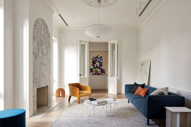 Bright, refined living room with white walls, ornate ceiling mouldings and ceiling roses, a sculptural marble fireplace, blue sofa, mustard accent chair, layered round coffee tables, and a neutral textured rug. Double doors lead to a hallway or adjoining room, with colourful artwork visible beyond.