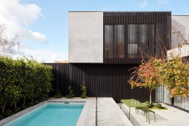 Contemporary two-storey residential facade viewed from a landscaped pool courtyard. The building combines smooth light rendered wall planes with a strongly contrasting black vertical slatted screen and matching dark lower fence, with glazing visible behind the screen. A narrow turquoise swimming pool, pale paved terrace, clipped hedge, and restrained outdoor furniture complete the composition. The image is bright and crisp with a modern, rectilinear architectural character.