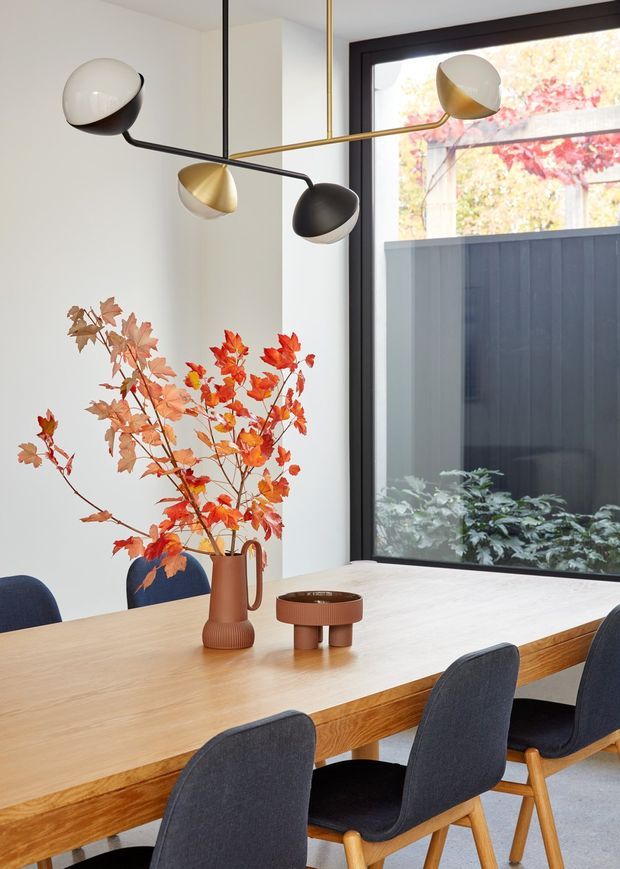A bright, contemporary dining area with a large light timber dining table surrounded by dark upholstered dining chairs. A sculptural multi-arm pendant light with black and brass arms hangs overhead. The room has white walls, dark-framed floor-to-ceiling glazing, and a simple ceramic vase with autumnal branches as a table centrepiece. A dark exterior fence and planting are visible through the window.