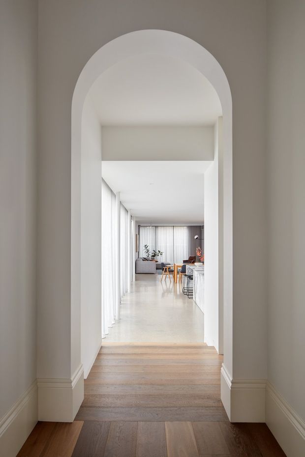 A view through a tall, rounded archway into a bright open-plan living and dining area. The foreground has dark timber flooring and detailed white skirting, transitioning to pale timber flooring in the adjoining space. Floor-to-ceiling sheer curtains line the left side, with a grey sofa, dining table and chairs, plants, and a light kitchen island visible in the distance. The interior is restrained, bright, and contemporary with strong architectural framing from the arched opening.
