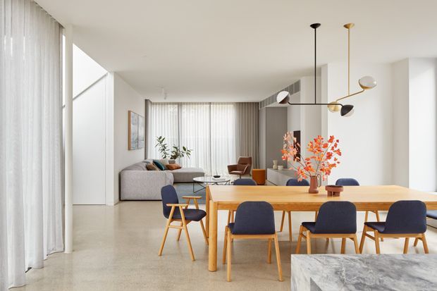 Bright open-plan contemporary living and dining area with floor-to-ceiling sheer curtains, pale polished concrete flooring, white walls, and an integrated kitchen island in the foreground. A light timber dining table with blue upholstered chairs occupies the main space, while a grey sofa and lounge seating sit in the background. A sculptural linear pendant light hangs above the dining area, with orange branches providing a small accent of colour.