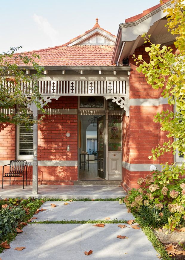 A sunlit front entrance to a heritage red-brick residence, featuring a terracotta tiled gable roof, ornate white timber fretwork and balustrades, decorative stained-glass entry doors, and a paved garden path bordered by mature planting. The facade has strong Victorian-era detailing and a welcoming, established garden setting.