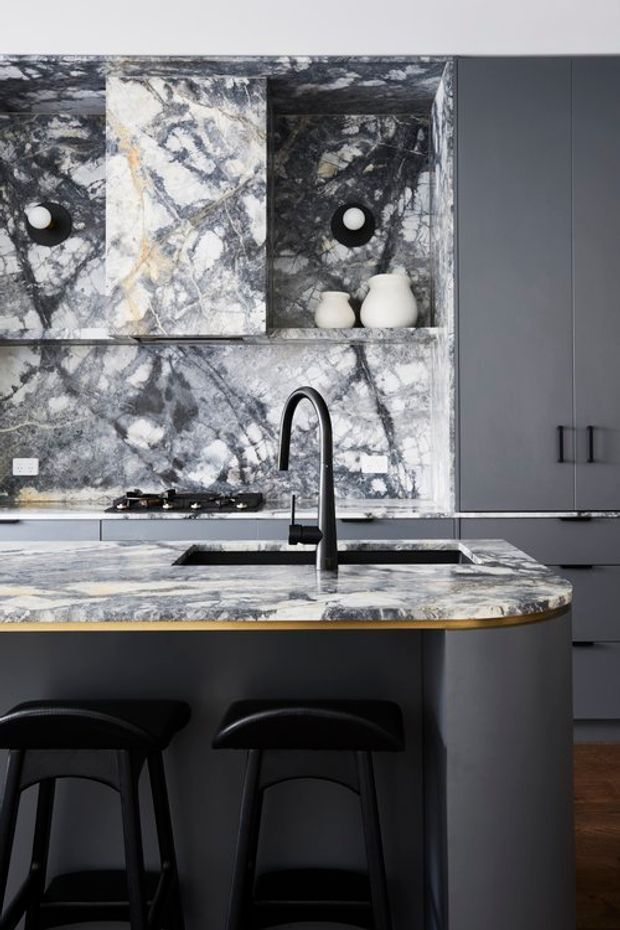 A contemporary kitchen centered on a large island with a dramatic veined stone benchtop and matching splashback. The island includes an undermount sink, tall black tap, rounded corner, and seating for two at black stools. Dark grey flat-panel cabinetry and black accents contrast with the pale grey, white, and charcoal stone. Open shelving displays simple white ceramic vessels, with warm timber flooring visible below.
