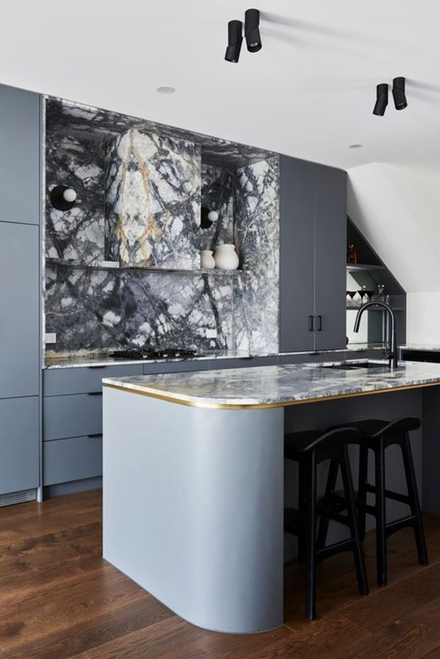 A sophisticated contemporary kitchen featuring dark grey-blue flat-panel cabinetry, a dramatic veined natural-stone backsplash and island benchtop, and a curved island end with a slim brass or gold trim. Black bar stools, a sculptural black tap, and black track lighting contrast with the pale cabinetry and warm timber flooring.