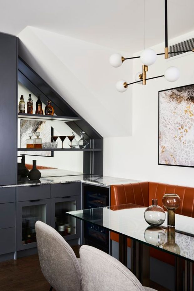 A sophisticated compact dining and entertaining area with a dark built-in bar and drinks cabinetry, mirrored shelving, wine storage, and a glossy stone counter. A glass-topped dining table with upholstered chairs sits beside a curved rust-orange banquette. The room features white walls, dark joinery, brass-accented globe pendant lighting, and framed abstract artwork.