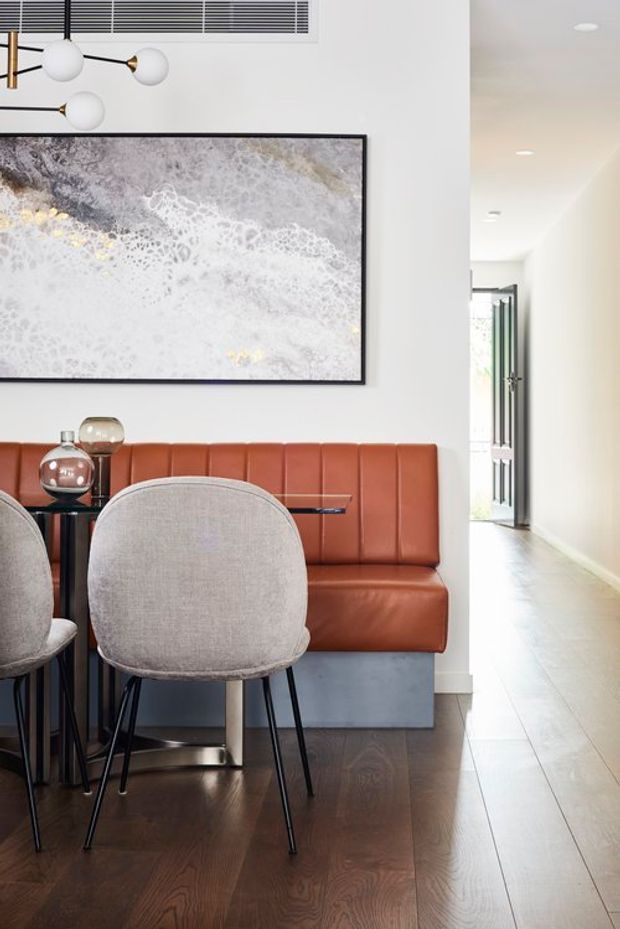 A bright, refined dining area with a built-in cognac leather banquette, round upholstered dining chairs, and a dark dining table on dark timber flooring. A large monochromatic abstract artwork and sculptural globe pendant add visual interest, while a light hallway extends to an open glazed entry door.