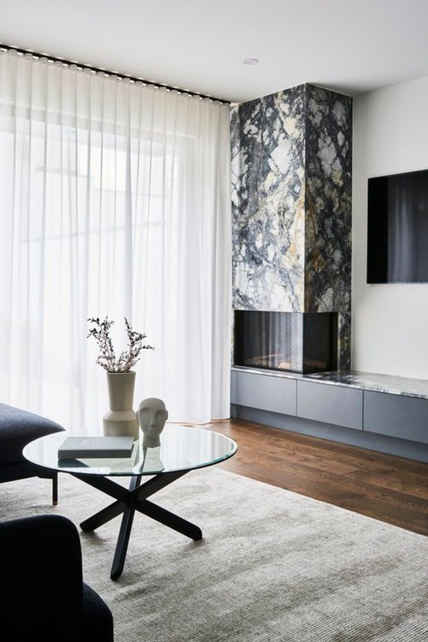 Contemporary living room with a dramatic floor-to-ceiling veined stone fireplace surround, built-in dark grey cabinetry and television, warm timber flooring, sheer full-height curtains, and a round glass coffee table with black sculptural legs. The room is bright and refined with a neutral palette and strong natural material contrast.