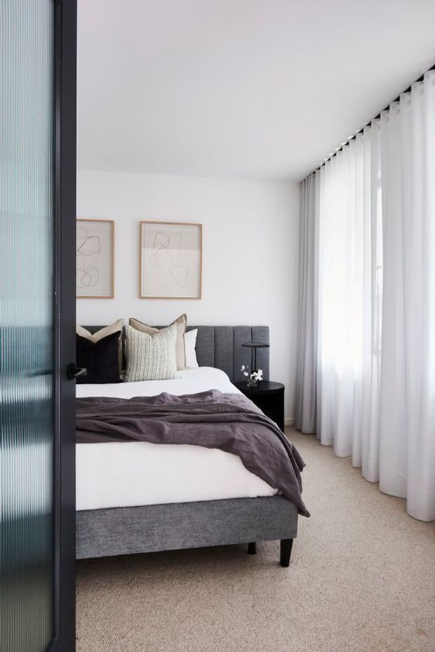 A bright, refined bedroom viewed through a dark-framed fluted-glass door. The room features a grey upholstered bed with white bedding and a deep mauve throw, layered neutral cushions, a black bedside table, minimalist framed artwork, full-height sheer curtains, and soft beige carpet. The palette is calm and predominantly white, grey, beige, and muted mauve.
