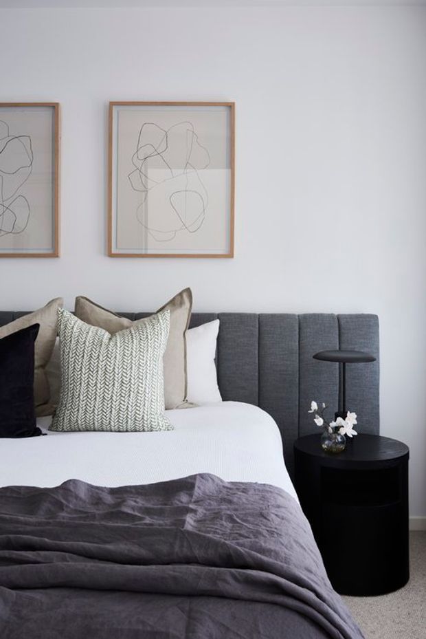 A calm, contemporary bedroom with a neatly made bed, charcoal upholstered vertical-panel headboard, layered white and grey bedding, textured neutral cushions, and a black round bedside table with a slender lamp and small floral arrangement. Two minimalist abstract artworks in light timber frames hang on the pale wall, while soft carpet and restrained monochromatic styling create a refined, serene atmosphere.
