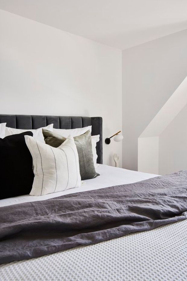 A minimalist bedroom with a neatly made bed, dark upholstered vertical-panel headboard, layered white and charcoal pillows, and a textured charcoal-grey throw. The room has white walls and ceiling, a simple black-and-white wall-mounted reading light, and a bright, restrained monochromatic palette.