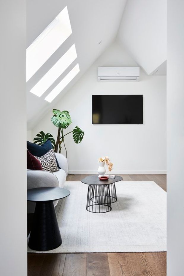 Bright, compact contemporary living room with a sloped ceiling and large skylights. The room features white walls, timber flooring, a wall-mounted television and split-system air conditioner, a grey sofa with patterned cushions, black wire-frame coffee tables, a large pale rug, and indoor tropical plants.