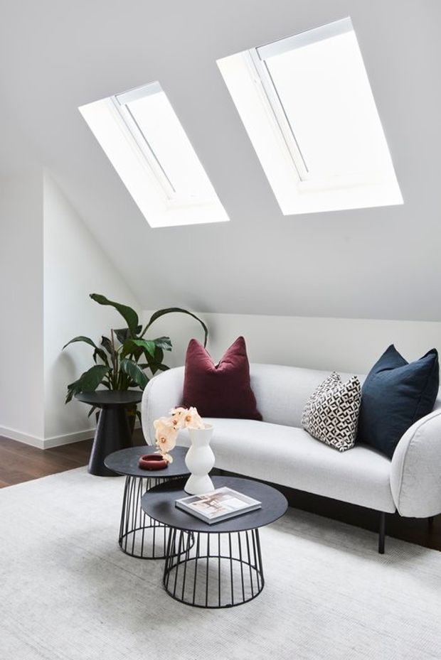 Bright contemporary living room in a compact attic or loft space, featuring a sloped white ceiling with two skylights, a light upholstered sofa, dark round nesting coffee tables, patterned and navy cushions, indoor greenery, and a pale area rug over dark timber flooring.