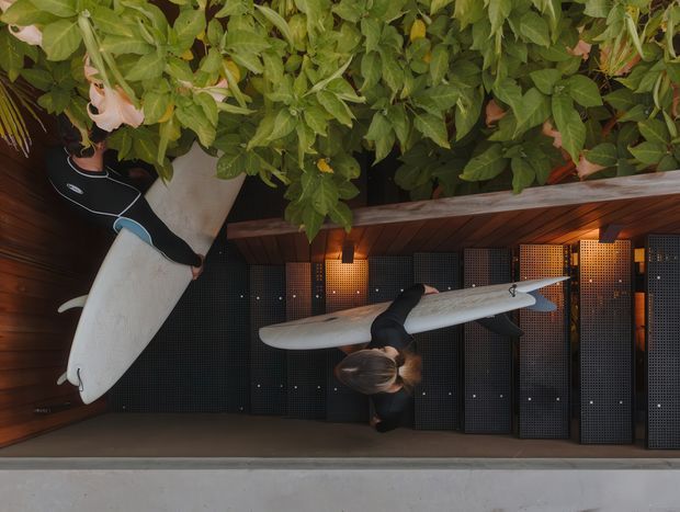 A small outdoor surfboard storage and changing area beneath dense greenery. Two surfboards are mounted against dark perforated locker panels, with warm timber soffit cladding, integrated lighting, and a concrete floor. A person is partially visible handling one of the boards.