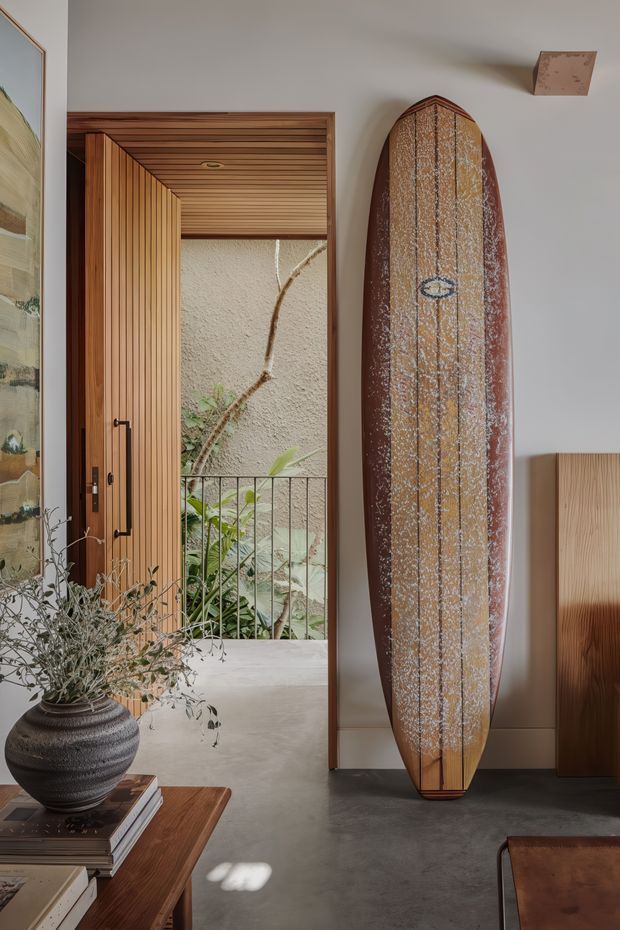 A warm, refined interior entry area with a tall timber pivot door opening to a planted courtyard or balcony. A vintage-style surfboard is displayed vertically against the wall, alongside timber cabinetry, a dark textured planter, books, and framed artwork. The space features extensive timber detailing around the doorway and ceiling, neutral plaster walls, and a smooth grey floor.