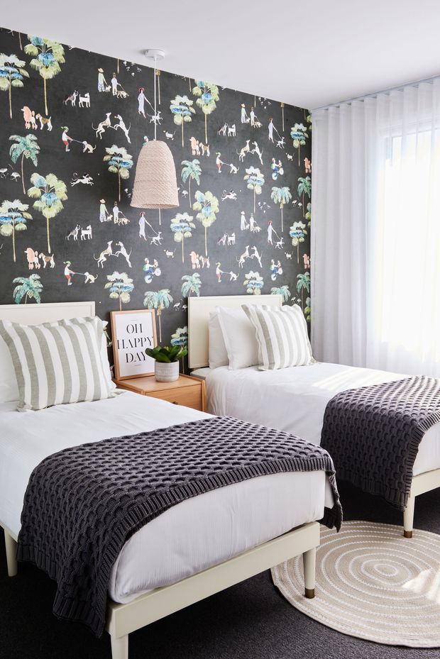 Bright twin bedroom with two single beds, white upholstered headboards and bedding, grey-and-white striped pillows, and dark charcoal knitted throws. A bold black patterned wallpaper featuring palm trees, animals, and figures forms the feature wall. A woven pendant light, small timber bedside table, framed artwork, indoor plant, sheer white curtains, and a round woven rug complete the playful yet refined interior.
