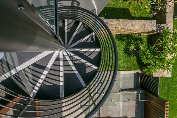 Overhead view of a modern spiral staircase with dark metal concentric railings and radial treads, adjacent to a contemporary landscaped courtyard with clipped lawn, stone paving, and architectural concrete and timber elements.