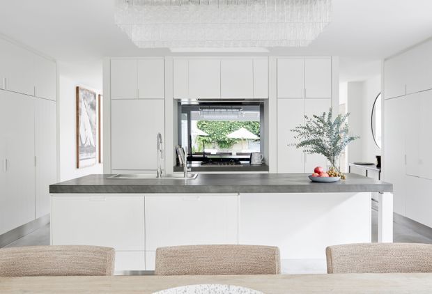 Bright, refined contemporary kitchen with extensive white full-height cabinetry, a large grey stone island with integrated sink, and a dining table with upholstered chairs in the foreground. A dark-framed window looks onto greenery, while a large linear crystal-style pendant adds a subtle luxurious accent.