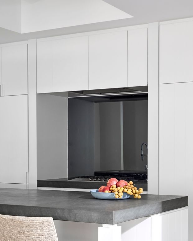 A minimalist contemporary kitchen with full-height white handleless cabinetry, a dark reflective splashback, integrated cooktop and rangehood, and a broad grey stone island benchtop. A bowl of fruit sits on the island, with a partially visible woven dining chair in the foreground. The space is bright, clean, and highly streamlined.
