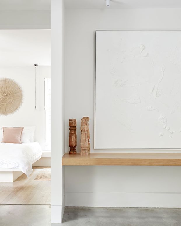 Bright, minimalist interior showing a bedroom partially visible through a wide opening on the left and a white built-in display ledge on the right. The space features white walls and ceiling, pale timber flooring and shelf, a smooth grey concrete floor in the foreground, simple timber decorative objects, and a large monochromatic textured artwork. Natural light creates an airy, calm atmosphere.