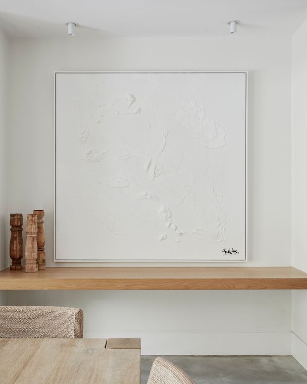 A minimalist light-toned interior vignette featuring a recessed wall shelf with a natural timber surface, a large monochromatic white textured artwork, and two carved wooden decorative objects. The lower portion shows a pale timber dining table, woven upholstered chairs, and a grey concrete-like floor. Small white directional ceiling spotlights illuminate the display.