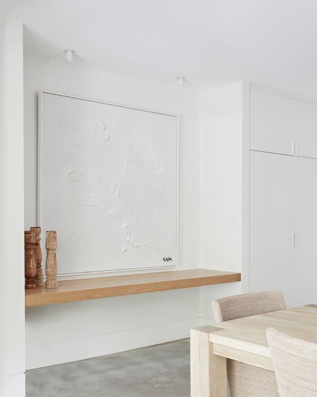 Bright, minimalist dining area with white walls and built-in cabinetry. A floating natural timber shelf displays a large monochromatic textured artwork and wooden sculptural objects, while a pale timber dining table with woven upholstered chairs occupies the foreground. The room has a polished concrete floor and recessed white ceiling spotlights.