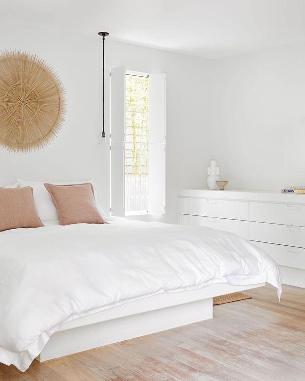 Bright, airy bedroom with a low white bed dressed in white bedding, soft blush-pink pillows, a large woven circular wall decoration, white built-in or freestanding drawer cabinetry, and pale timber flooring. A tall narrow window brings in natural light, while a slim black pendant adds contrast.