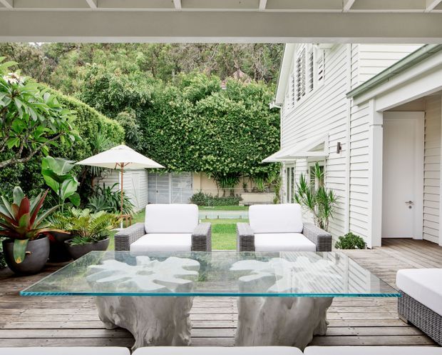 A refined outdoor entertaining area viewed from beneath a covered structure, with a timber deck, glass-topped sculptural coffee table, white outdoor lounge seating, and lush tropical planting. A white weatherboard-clad house with louvred windows and an open doorway borders the space, while a manicured courtyard and umbrella are visible beyond.