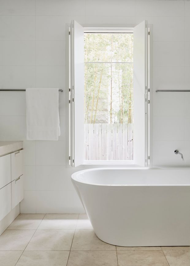 Bright minimalist bathroom featuring a freestanding white bathtub beneath an open casement window, white tiled walls, pale tiled flooring, simple white cabinetry, and understated chrome towel and tap fixtures. Natural greenery is visible outside the window.
