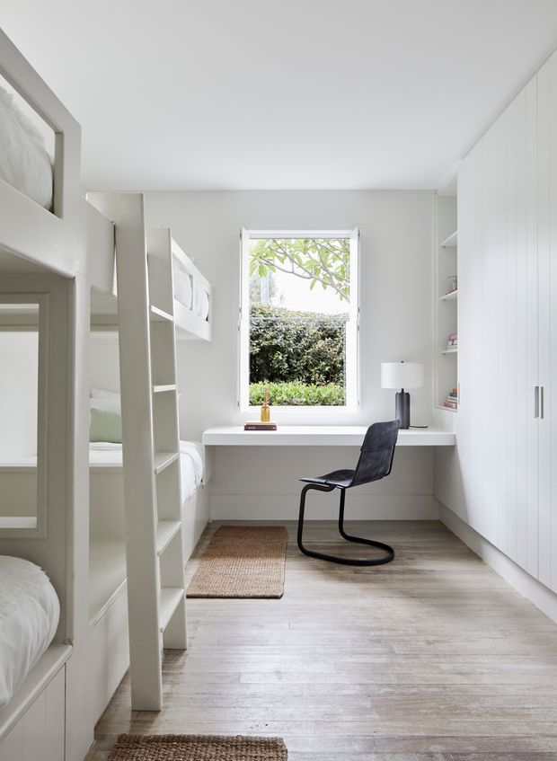Bright, compact white bedroom with built-in bunk beds along the left wall, a built-in study desk beneath a window, a black rocking chair, full-height white storage cabinetry, pale timber flooring, and woven rugs. The space has a clean, contemporary minimalist feel with abundant natural light and garden views.