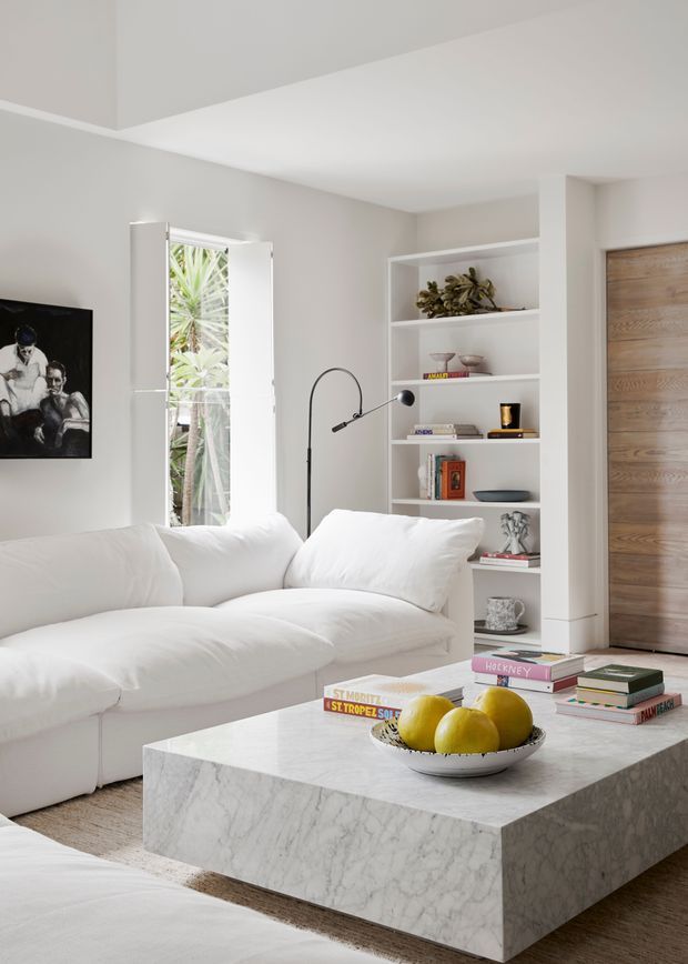 Bright, refined living room featuring a large white upholstered sectional, a substantial veined marble coffee table, built-in white shelving with books and objects, a slim black arched floor lamp, and a vertical timber door. Neutral plaster walls, white shutters, and abundant daylight create a clean contemporary interior.