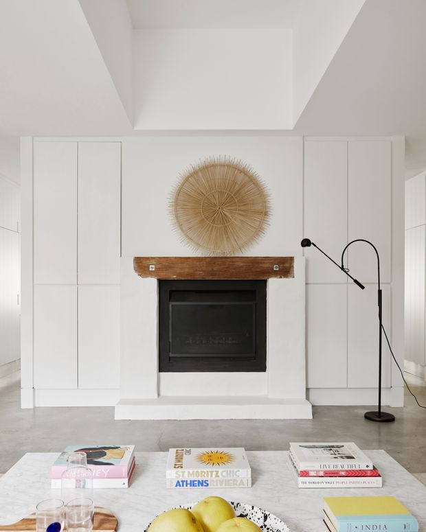 Bright minimalist living room centred on a black fireplace with a rustic timber mantel, built-in white cabinetry, and a large woven sunburst wall feature. A low marble or stone coffee table with books, glasses, and fruit occupies the foreground. The room has a pale polished concrete floor, white walls and joinery, and a sculptural black floor lamp.