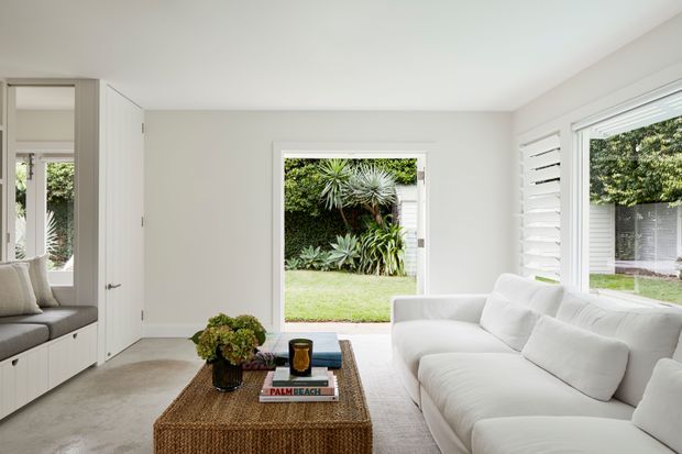 Bright, refined living room with a white slipcovered sectional sofa, built-in window seat and cabinetry, pale neutral walls, large windows with white plantation shutters, and a woven rectangular coffee table. The room opens directly onto a lush landscaped garden, creating an airy indoor-outdoor connection.