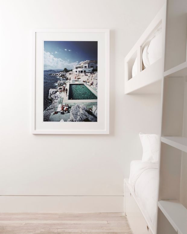 A bright, minimalist bedroom corner with white built-in bunk-style sleeping cabinetry on the right, white bedding, pale timber flooring, and a framed coastal photograph on a soft white wall.