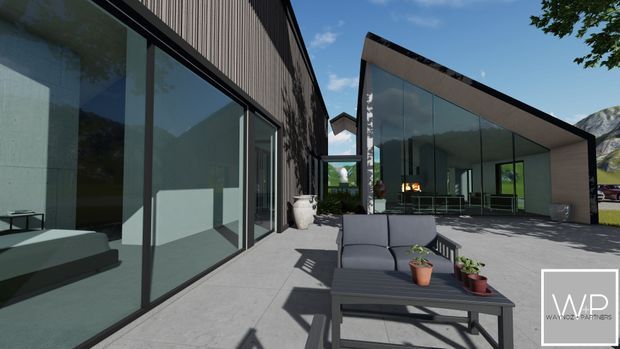 Contemporary architectural exterior viewed from a paved outdoor terrace, with dark vertical cladding, expansive floor-to-ceiling glazing, and a sharply angled mono-pitch roof volume. Outdoor lounge furniture occupies the foreground, while a bright glazed interior is visible through the facade. Mountainous surroundings are reflected in the glass and visible at the edges.