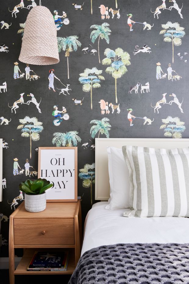 A styled bedroom corner featuring a white bed with a grey-and-white striped cushion and chunky knitted throw, a mid-century-inspired timber bedside table, potted plant, and framed typographic artwork. The dark feature wall is covered in playful illustrated wallpaper depicting palm trees, animals, and figures, with a large woven pendant light hanging above.