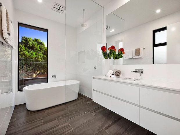 Bright contemporary bathroom with a freestanding white bathtub, frameless glass shower enclosure, large mirrored wall, floating white vanity with integrated basin, and dark wood-look tiled flooring. Black-framed windows bring in natural light and overlook greenery. Red flowers and neatly folded towels provide small decorative accents.