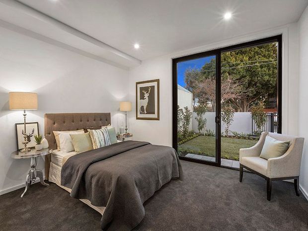 A bright, contemporary bedroom with a tufted upholstered bed, layered neutral bedding, dark grey carpet, white walls and recessed ceiling lights. A large black-framed sliding glass door opens to a small landscaped courtyard, while a framed artwork, bedside tables and an upholstered armchair complete the room.