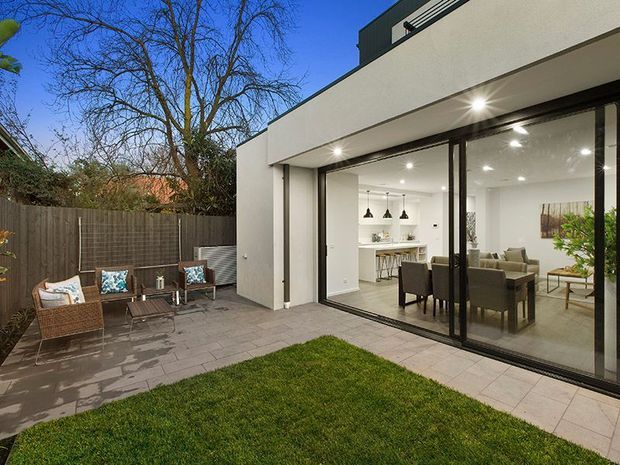 Contemporary two-storey home viewed from a landscaped courtyard, with a white rendered facade, broad black-framed floor-to-ceiling glazing, and a covered outdoor terrace. Through the glazing, a bright open-plan dining and living area with a compact kitchen is visible. The courtyard includes pale rectangular paving, lawn, a timber privacy fence, and an outdoor seating area.