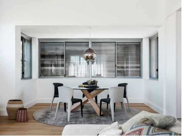 Bright, contemporary dining area in a white-lined bay-window setting. A rectangular glass dining table with crossed timber legs is surrounded by mixed black and white chairs, positioned on a round grey rug over timber flooring. Horizontal blinds cover the surrounding windows, while a reflective spherical pendant hangs centrally above the table. Part of a pale upholstered sofa with patterned cushions is visible in the foreground.