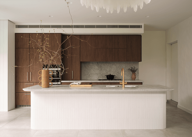 A refined contemporary kitchen with a large rounded island clad in white vertical ribbed tiles, a pale stone benchtop, dark timber cabinetry, and a matching stone splashback. Brass tapware, integrated appliances, open shelving, and sculptural pendant lighting add warm detail to the otherwise minimalist space.