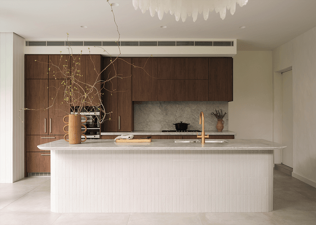 A refined contemporary kitchen viewed straight on, centred on a large island with a vertically fluted white finish and a pale stone benchtop. The rear wall features extensive dark timber cabinetry, integrated appliances, a mottled stone splashback, and a brass-toned tap. Neutral tiled flooring, white walls, concealed ventilation, and a sculptural pendant light complete the calm, warm-toned interior.