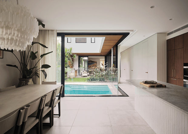 A refined open-plan kitchen and dining area with a long stone island, integrated pale cabinetry, dark timber kitchen joinery, and a dining table beneath a sculptural pendant light. Full-height glazing opens toward an internal courtyard and swimming pool, while timber ceiling lining and abundant planting add warmth and an indoor-outdoor connection.