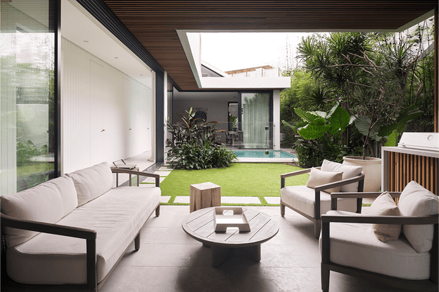 A refined contemporary covered outdoor lounge opening onto a landscaped courtyard and compact pool. The space features a large cream upholstered sofa and matching armchairs, a round low coffee table, pale stone or concrete flooring, dark-framed full-height glazing, and a dramatic timber-lined slatted ceiling with a large rectangular skylight opening. Lush tropical planting surrounds the courtyard, with an outdoor kitchen or barbecue unit visible at right.
