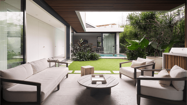 A refined contemporary outdoor lounge arranged around a low circular coffee table, with a large upholstered sofa and matching armchairs beneath a dark timber-lined covered ceiling. The space opens onto a manicured courtyard with clipped lawn, tropical planting, a narrow swimming pool, and glazed modern architectural forms. The palette is neutral and warm, combining pale upholstery, dark frames, timber soffits, and lush green landscaping.