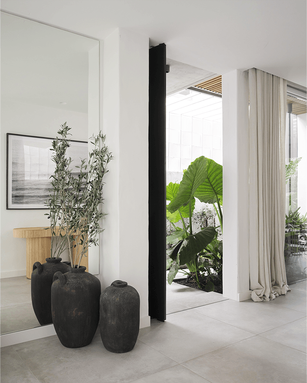 Bright minimalist interior with large pale stone or porcelain floor tiles, white walls, full-height glazing, and a dramatic black pivot-style door frame. A full-height mirror reflects a console and artwork, while large dark sculptural vases with leafy branches create a natural focal point. Floor-to-ceiling sheer curtains open toward a lush planted courtyard, and a timber slatted ceiling detail is visible near the glazing.
