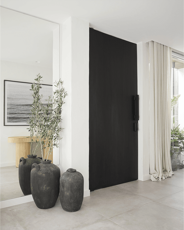 Minimalist interior entryway featuring a tall matte-black pivot door set within white walls, large light-grey floor tiles, full-height cream curtains, a full-height mirror, and sculptural dark ceramic planters with olive branches. A coastal artwork and timber console are visible in the adjacent reflected space.